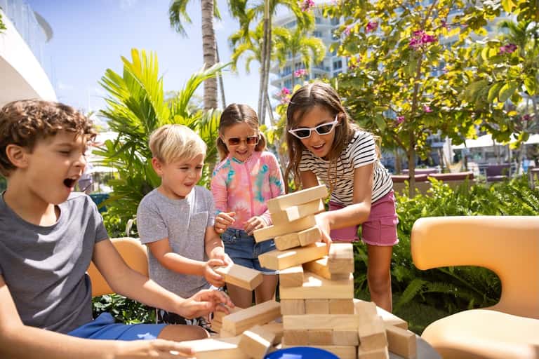 Children playing with wooden blocks outdoors under palm trees and flowering vines at resort