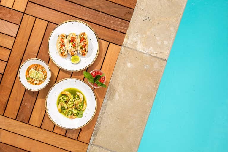 Gourmet dishes on wooden deck overlooking bright turquoise swimming pool
