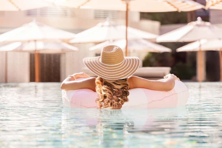 Woman floating in pool wearing wide-brimmed hat with white umbrellas and loungers visible