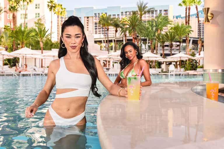 Two women enjoying resort pool with in-pool bar, tropical palms, and resort buildings in background