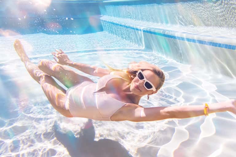 Woman floating underwater in swimming pool wearing white swimsuit and sunglasses