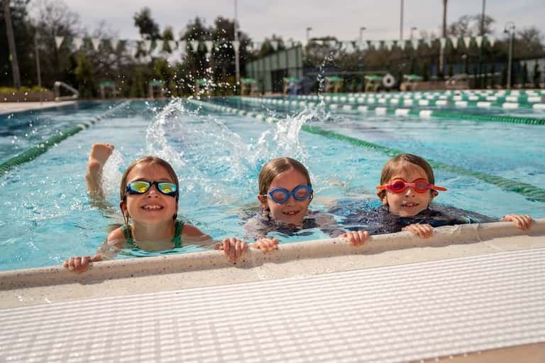 Three young children swimming in lap pool wearing colorful goggles, smiling at camera poolside