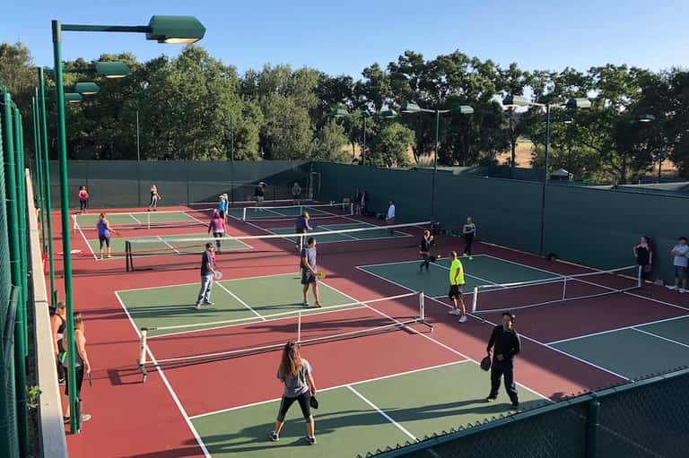 Outdoor tennis courts with multiple players, red and green surfaces, surrounded by green boundary fence
