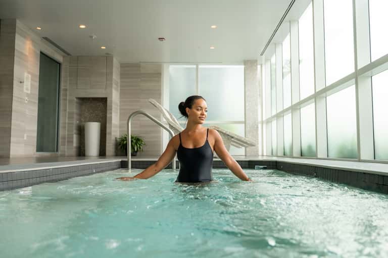 Woman relaxing in modern spa tub with floor-to-ceiling windows and neutral tile design
