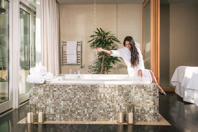 Woman in white robe arranging flowers beside luxurious bathroom tub with tile surround