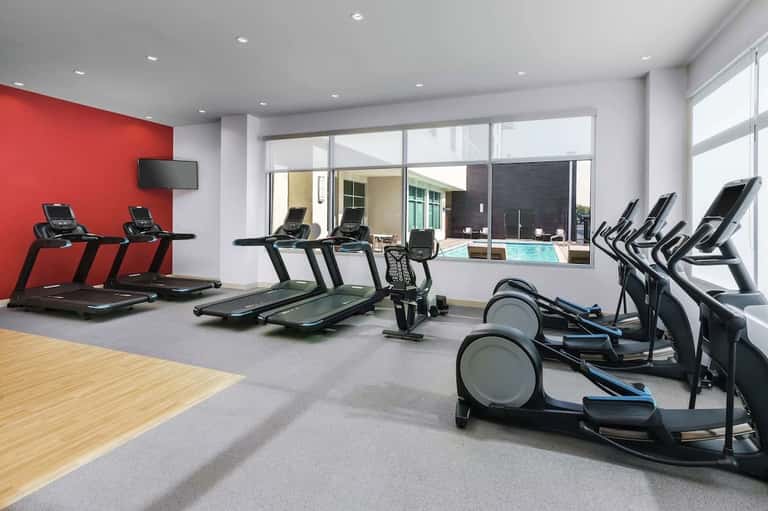 Bright fitness center with treadmills, exercise bikes, red accent wall, and pool view through large windows