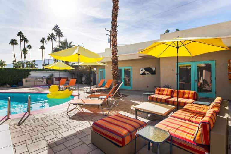 Resort pool area with yellow umbrellas, colorful lounge chairs, palm trees, and modern turquoise building