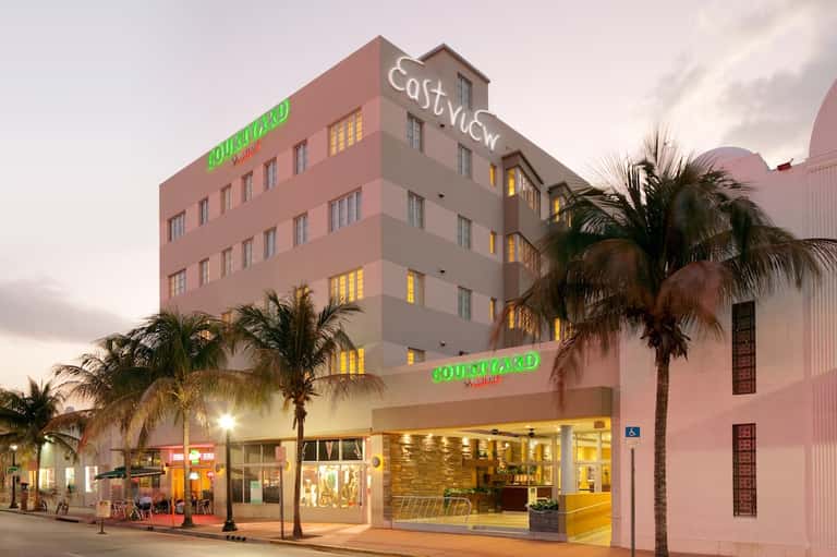 Pink art deco hotel building illuminated at dusk with palm trees and street-level retail