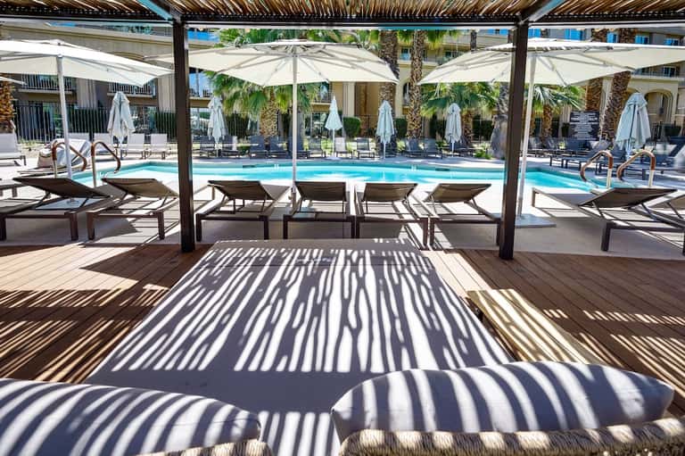 Resort pool deck with lounge chairs under pergola shade, white umbrellas, and palm trees with bright blue pool water