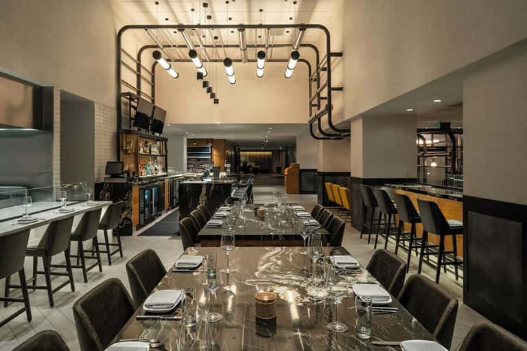 Contemporary hotel restaurant and bar with marble tables, geometric ceiling fixtures, and open kitchen