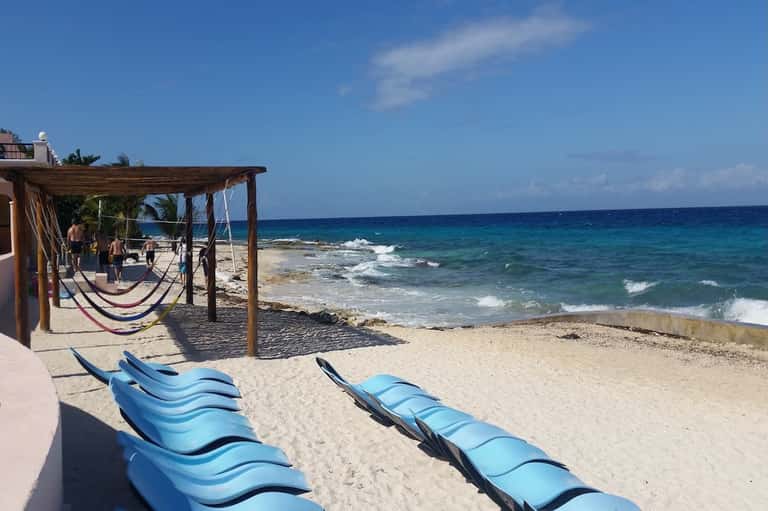 Beach scene with blue lounge chairs, wooden pergola, hammocks, and ocean waves with palm trees