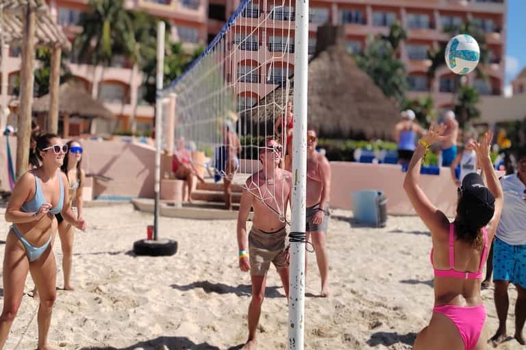 Beach volleyball game at resort with players in action, sand court, palm trees, and pink buildings in background