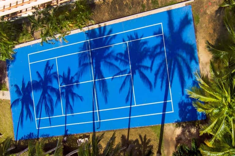 Aerial view of bright blue tennis courts surrounded by palm trees casting shadows