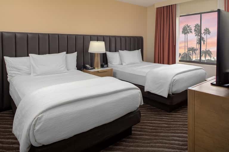 Hotel guest room with two queen beds, gray headboards, warm lighting, and tropical sunset view