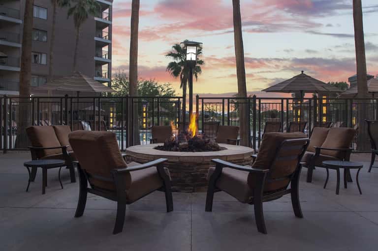 Outdoor fire pit lounge at dusk with brown cushioned chairs, palm trees, and sunset sky with building in background.