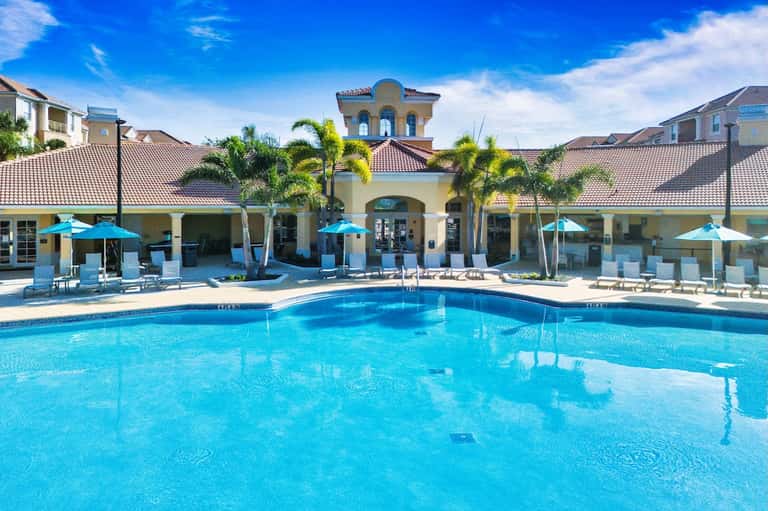 Resort pool with bright blue water, palm trees, blue umbrellas, and Spanish-style building with dome entrance