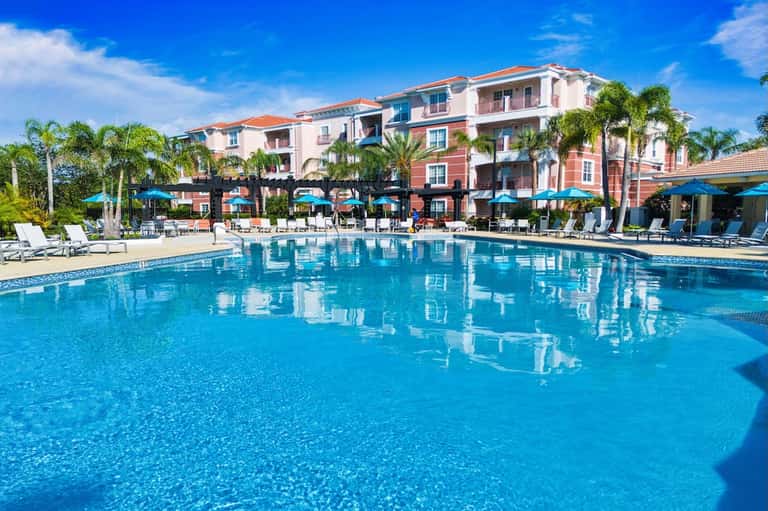 Resort pool with colorful building, palm trees, blue umbrellas, and lounging guests