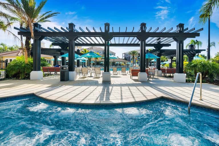 Outdoor hot tub and spa area under black pergola with palm trees, turquoise umbrellas, and resort in background