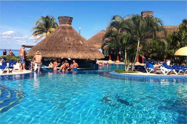 Resort pool with thatched-roof bar, tropical palm trees, lounge chairs, and guests enjoying swimming