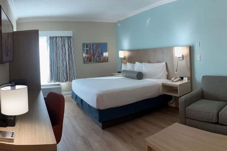 Modern hotel bedroom with queen bed, turquoise accent wall, desk, and sitting area