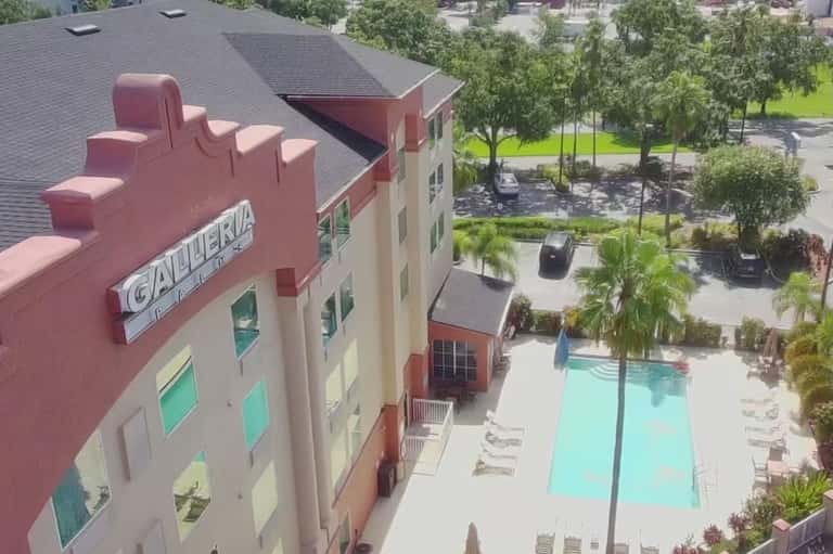 Aerial view of residential complex with colorful building, pool, green landscaping, and surrounding development