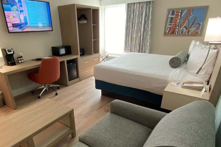 Modern hotel room with queen bed, wood desk, rust accent chair, flat-screen TV, and neutral color palette