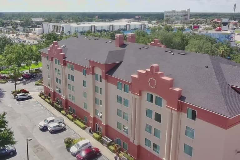 Aerial view of multi-story hotel with red and cream facade, flat roof, parking lot, and surrounding cityscape