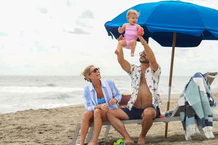 Family enjoying beach time under blue umbrella with toddler, ocean waves visible in background.