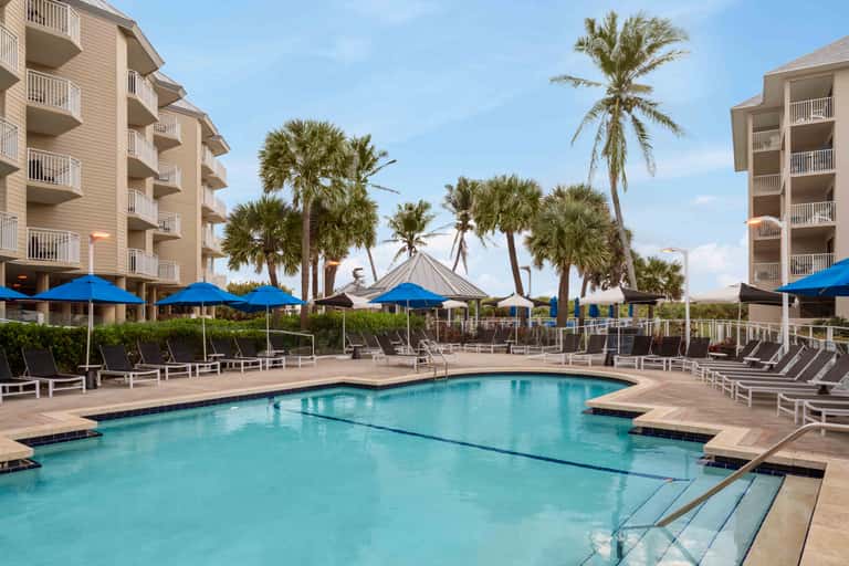 Resort pool area with blue umbrellas, lounge chairs, palm trees, and residential buildings