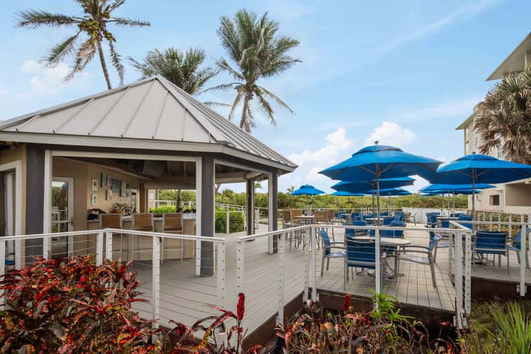 Beachfront gazebo and deck with blue umbrellas, lounge chairs, palm trees, and coastal views