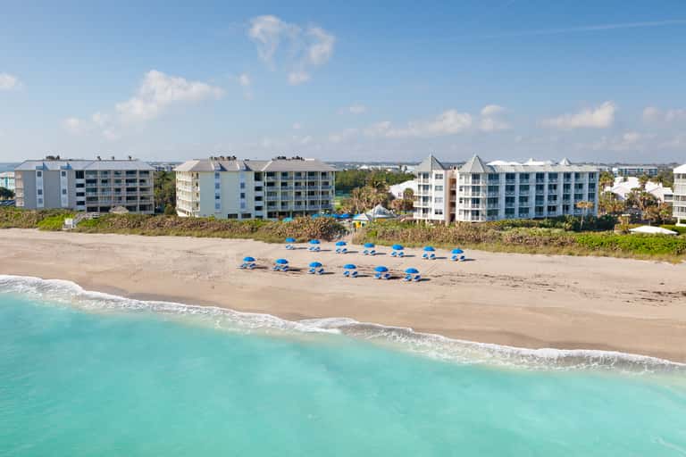 Beachfront resort with multiple pastel-colored buildings and blue umbrellas on sandy beach