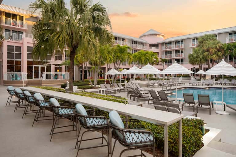 Resort pool area at sunset with pink buildings, palm trees, white umbrellas, and lounge seating
