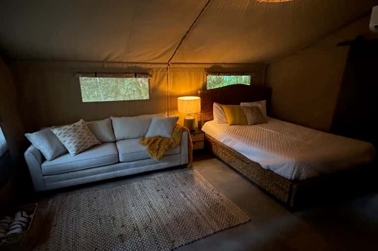 Glamping tent interior with queen bed, sofa, warm lighting, and windows to nature