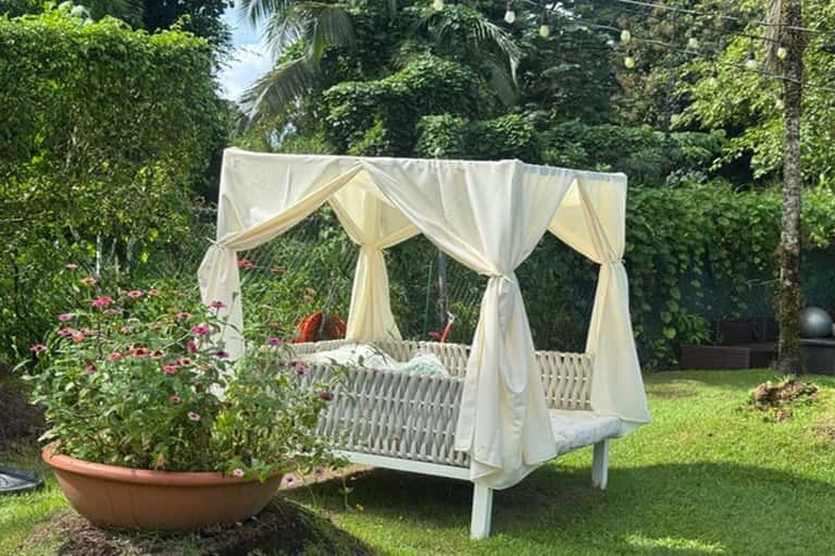 White canopy bed with cream drapes in lush garden setting with flowering plants and tropical greenery