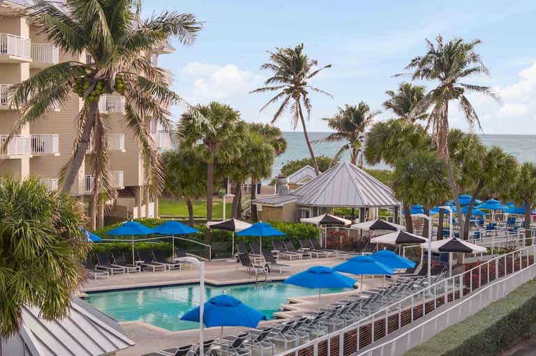 Resort pool area surrounded by palm trees, blue umbrellas, and white gazebos with ocean views
