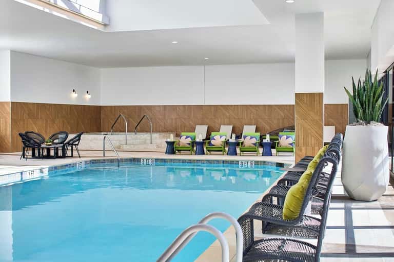 Modern indoor pool with high ceilings, wooden wall accents, green lounge chairs, black wicker seating, and skylights