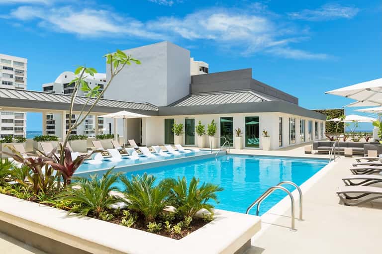 Resort pool and terrace with white umbrellas, tropical landscaping, and ocean view under blue sky
