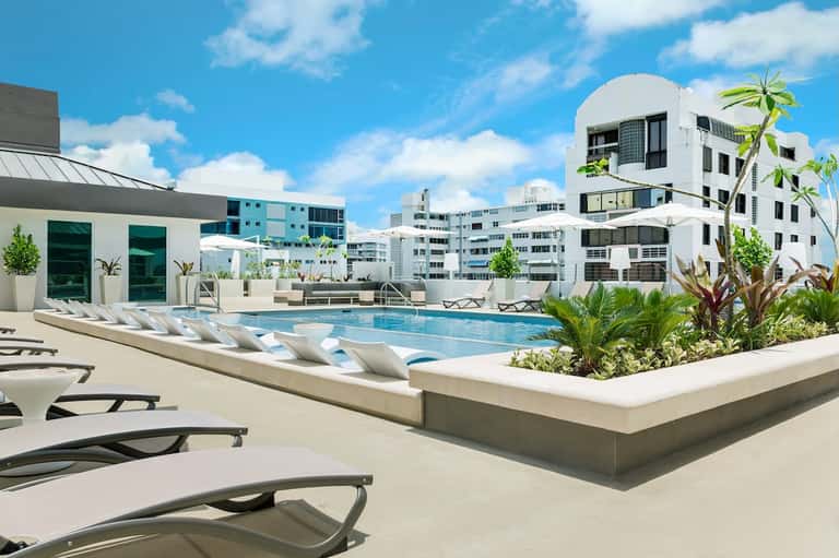 Resort pool with white loungers, tropical plants, modern white buildings, and clear blue sky