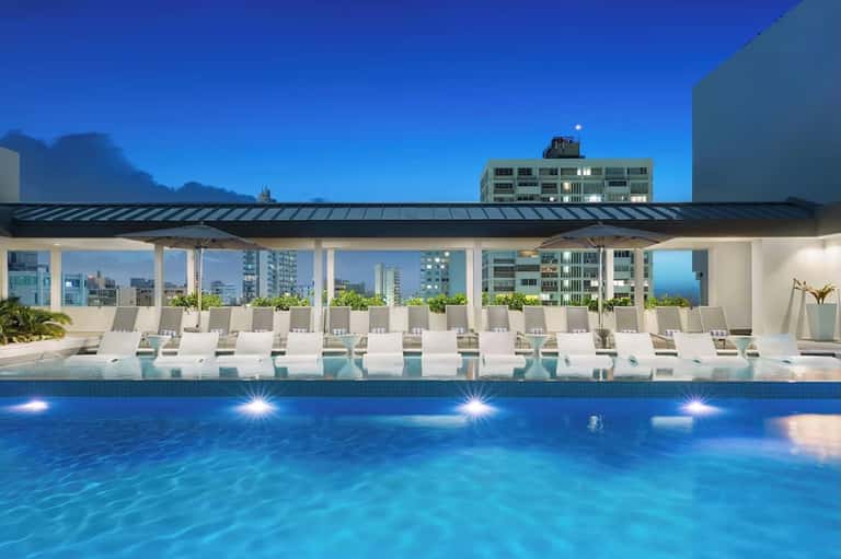 Rooftop pool deck at night with white loungers, pergola, city views, and ambient lighting