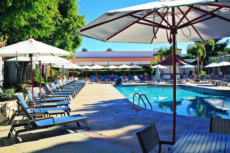Resort pool area with blue lounge chairs, white umbrellas, and lush green landscaping under clear sky