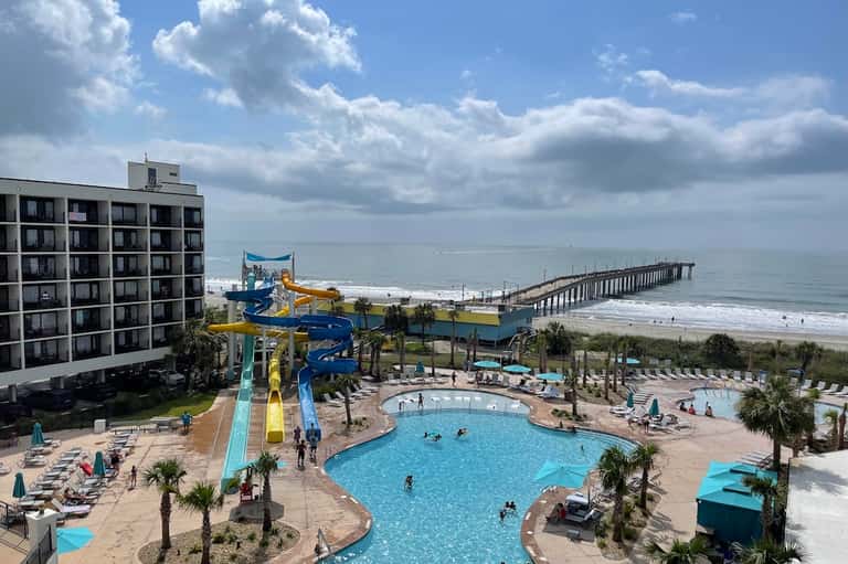 Beachfront resort with large pool, water slide, hotel tower, and ocean pier visible