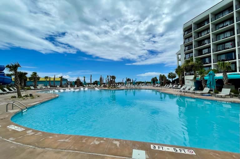 Large resort pool with cabanas, palm trees, multi-level building, and blue sky backdrop