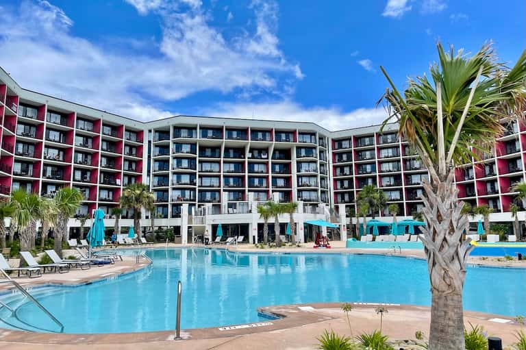 Large resort pool with blue water, modern multi-story hotel building, lounge chairs, and turquoise umbrellas