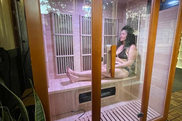 Infrared sauna interior with woman sitting on wooden bench, glass door, and purple lighting