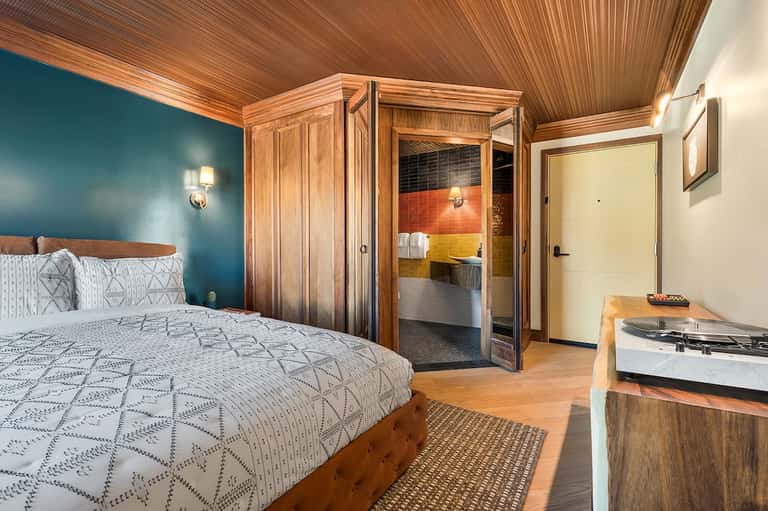 Modern bedroom with teal accent wall, wooden furnishings, and colorful tiled bathroom visible