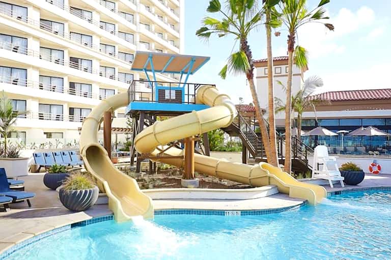 Resort pool with yellow waterslide, palm trees, and multi-story hotel building in background