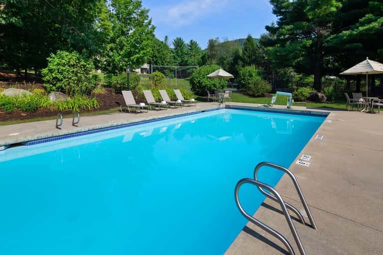 Resort swimming pool surrounded by lounge chairs, umbrellas, and tree-lined landscaping on sunny day