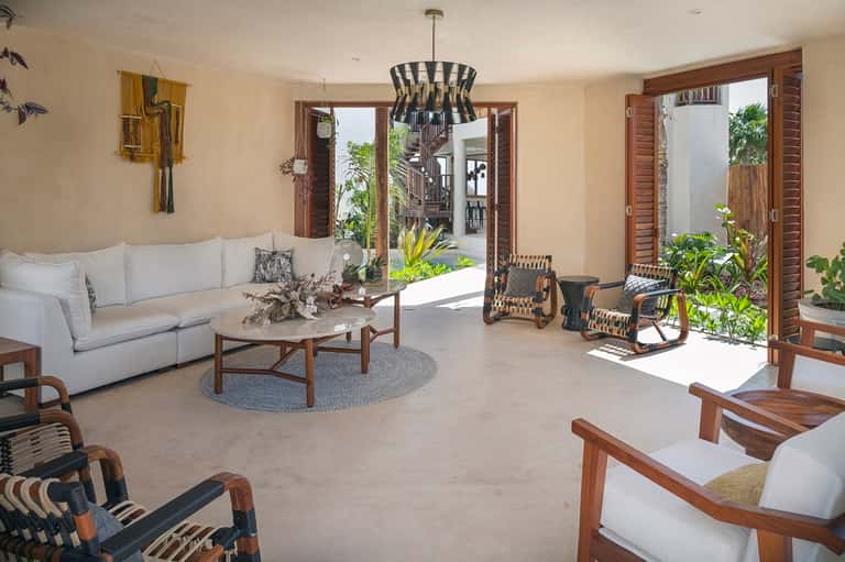 Bright open-concept living room with cream sofa, wooden furnishings, large windows opening to tropical courtyard.