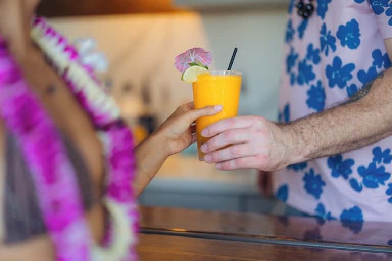 Hands exchanging a tropical yellow cocktail with pineapple garnish and decorative lei background