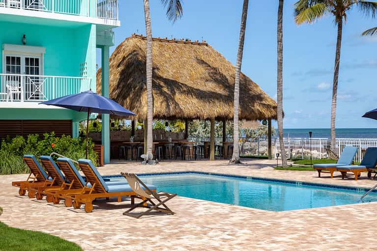 Beachfront resort pool with lounge chairs, thatched-roof bar, palm trees, and ocean view